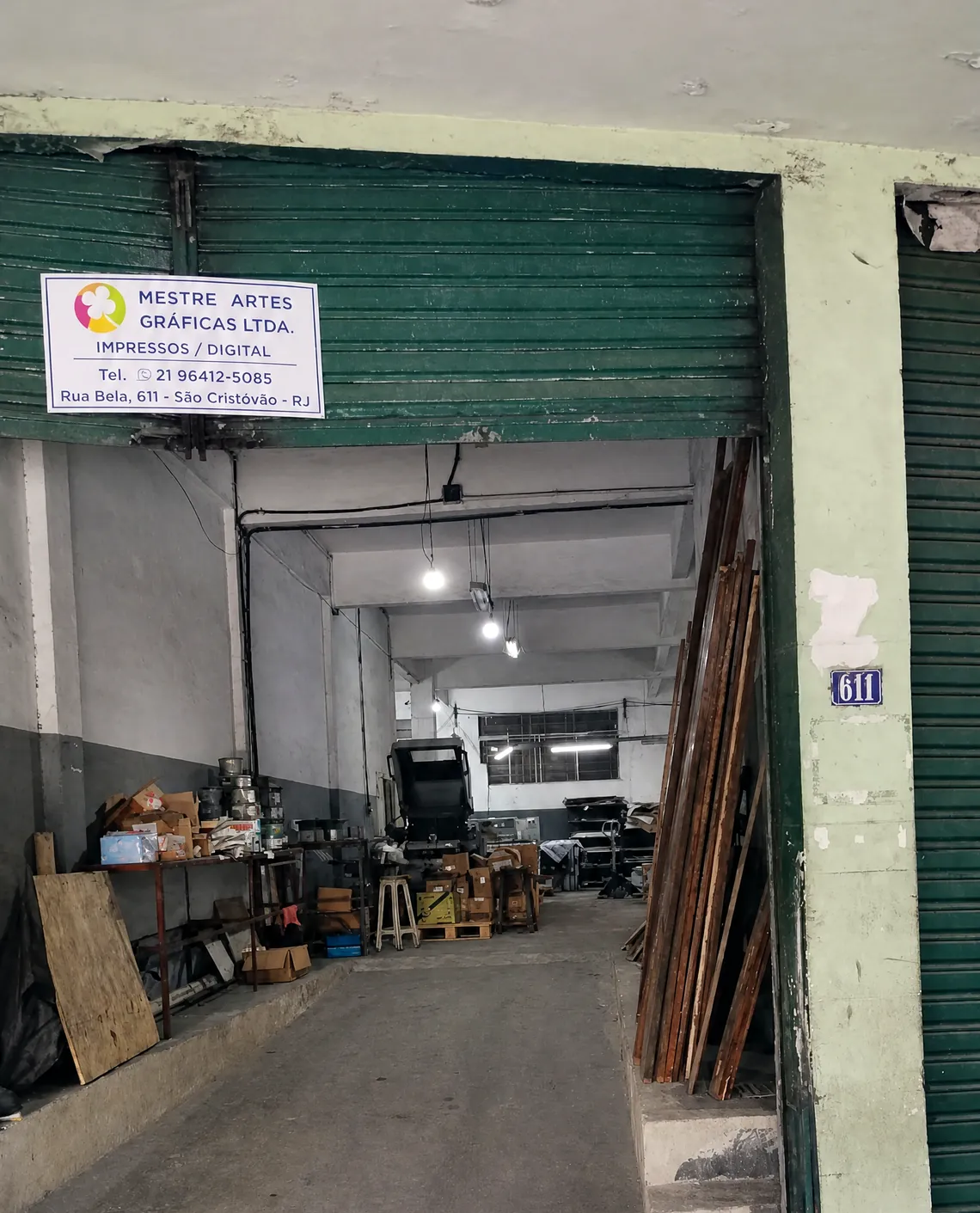 Entrada da Mestre Gráfica na Rua Bela, 611, em São Cristóvão: a placa da empresa sobre o portão verde, o número 611 na parede e a oficina ao fundo